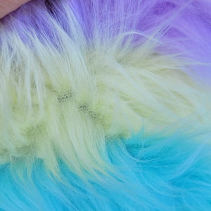 May include: Close-up of a multi-coloured faux fur fabric. The fabric is a gradient of purple, yellow, and blue.