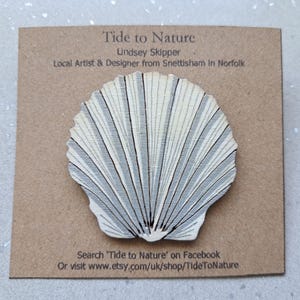 May include: A wooden seashell ornament painted in white and grey with a textured surface. The ornament is on a brown card with the text "Tide to Nature" and "Lindsey Skipper" printed on it.