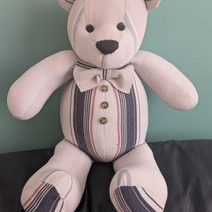 Op de afbeelding: Een knuffelbeer gemaakt van beige stof met donkerblauwe en rode strepen. De beer heeft een bijpassende vlinderdas bij de nek, drie bruine knopen in het midden van de romp, zwarte knoopogen en een zwarte stoffen neus.