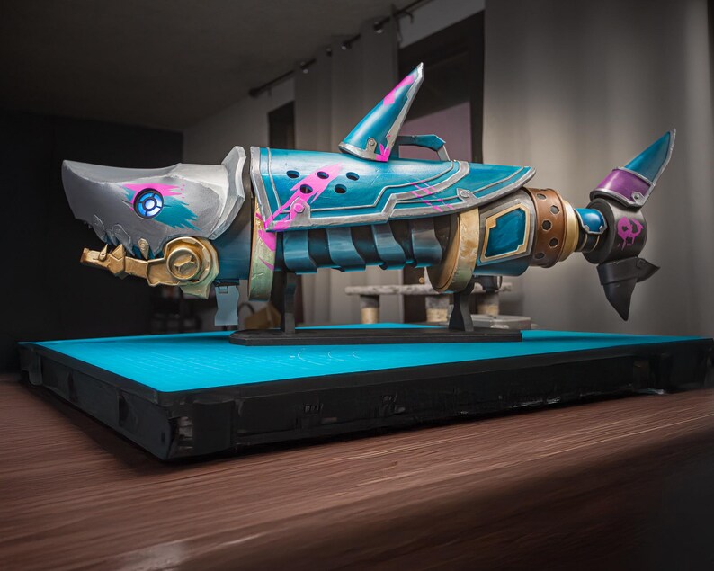 K&ouml;nnte beinhalten: Stilisierte, detaillierte Hai-Skulptur in Blau-, Silber- und Goldt&ouml;nen mit rosa Akzenten. Die Skulptur weist mechanische Details auf und ist auf einem blauen Sockel montiert. Die Schwanzflosse des Hais hat ein Totenkopf-Design.