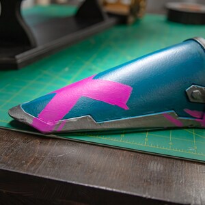 K&ouml;nnte beinhalten: Ein zylindrischer Armschutz in T&uuml;rkis mit einem rosa X-f&ouml;rmigen Design. Die Kanten sind silbern umrandet. Der Armschutz liegt auf einer gr&uuml;nen Schneidematte und einer Holzoberfl&auml;che.