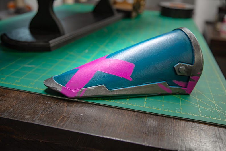 K&ouml;nnte beinhalten: Ein zylindrischer Armschutz in T&uuml;rkis mit einem rosa X-f&ouml;rmigen Design. Die Kanten sind silbern umrandet. Der Armschutz liegt auf einer gr&uuml;nen Schneidematte und einer Holzoberfl&auml;che.