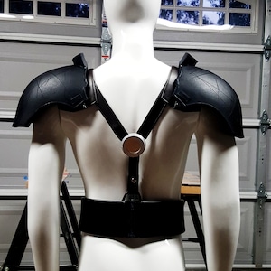 May include: A white mannequin wearing a black leather harness with shoulder pads. The harness has a circular metal buckle in the back.