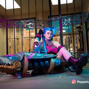 K&ouml;nnte beinhalten: Ein Cosplay-Requisit in Form einer haif&ouml;rmigen Waffe, haupts&auml;chlich blau und gold, mit rosa Akzenten. Das Requisit weist mechanische Details und einen haifischartigen Mund auf. Der Hintergrund zeigt ein Metalltor und ein Geb&auml;ude.
