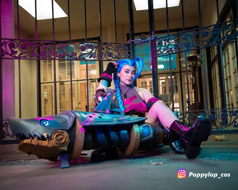 K&ouml;nnte beinhalten: Ein Cosplay-Requisit in Form einer haif&ouml;rmigen Waffe, haupts&auml;chlich blau und gold, mit rosa Akzenten. Das Requisit weist mechanische Details und einen haifischartigen Mund auf. Der Hintergrund zeigt ein Metalltor und ein Geb&auml;ude.