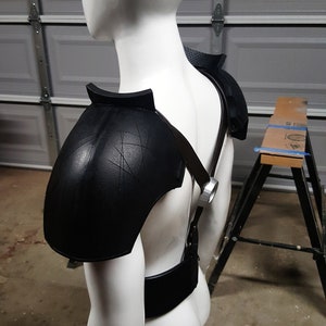 May include: A white mannequin wearing a black leather shoulder armor with straps and a black belt.