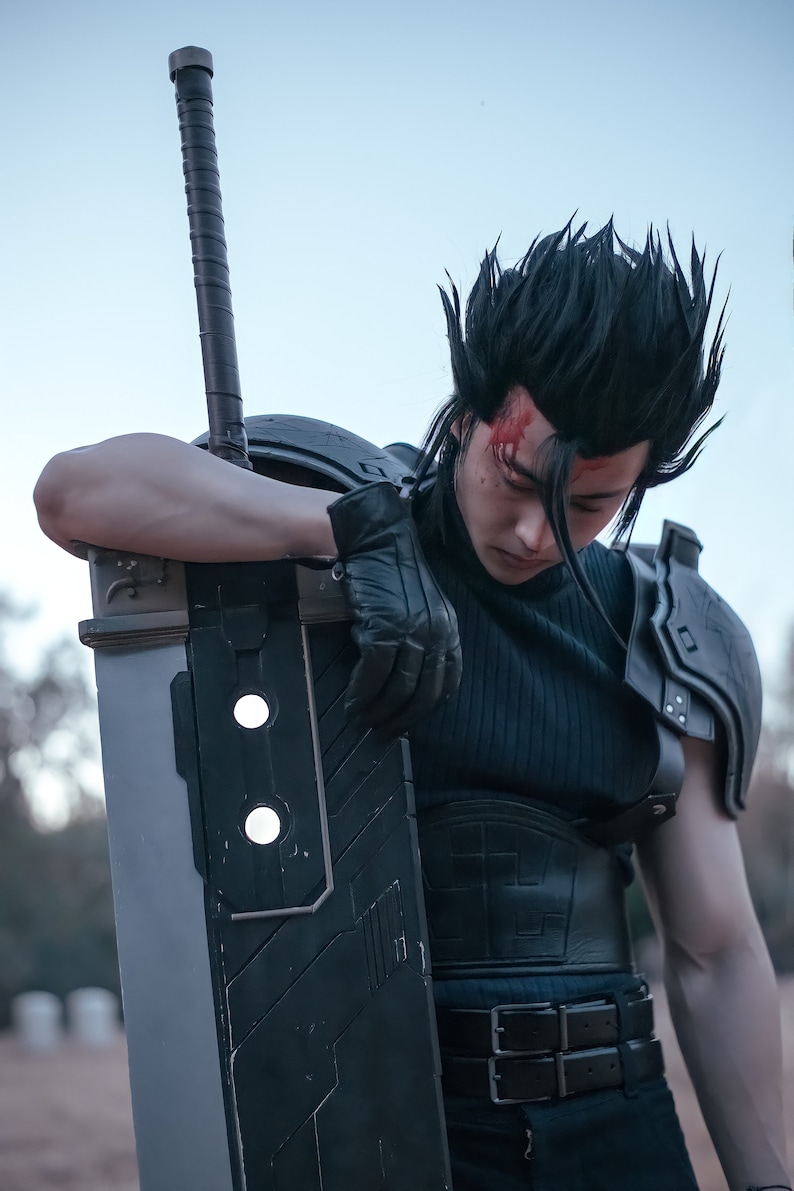 May include: A person dressed as a video game character is holding a large, silver sword with a black handle. The character is wearing a black shirt, black pants, and a black belt with two buckles. The character has black hair and a red mark on their forehead.