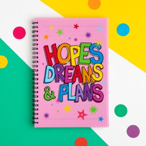 May include: A pink spiral-bound notebook featuring the text "HOPES DREAMS & PLANS" in vibrant, multi-coloured lettering. The cover is adorned with star motifs. The background is a mix of green, yellow, and white with colourful polka dots.