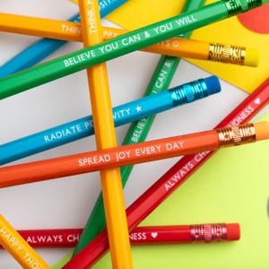 Pu&ograve; includere: Primo piano di matite colorate con frasi motivazionali in testo bianco. Le matite sono di vari colori, tra cui giallo, verde, blu, arancione e rosso. Ogni matita ha una ghiera dorata e una gomma rosa o blu.