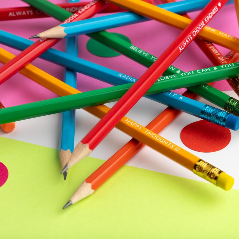 Pu&ograve; includere: Un assortimento di matite colorate con frasi motivazionali stampate, come "Always Choose Kindness" e "Happy Thoughts". Le matite, in vari colori tra cui rosso, blu, verde e giallo, sono disposte su uno sfondo rosa e verde a pois.