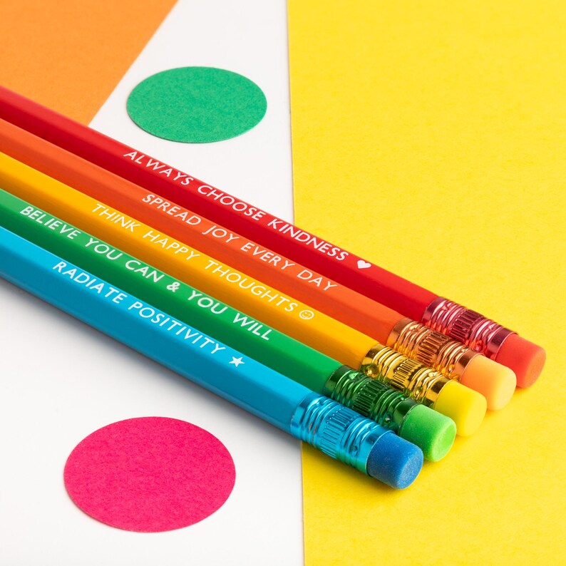 Pu&ograve; includere: Un set di sei matite arcobaleno con frasi motivazionali stampate in bianco. Le matite sono disposte diagonalmente su uno sfondo di carta gialla, bianca e arancione con accenti circolari colorati. Ogni matita ha una gomma di colore diverso.