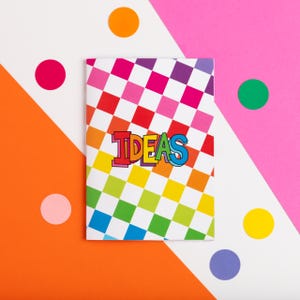 May include: A notebook with a rainbow checkered pattern and the word "IDEAS" in colorful block letters. The notebook is on a white background with orange, pink, and white geometric shapes and colorful polka dots.