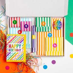May include: A colourful birthday gift box with a card that says "Happy Birthday" and a rainbow striped paper bag with the number "7" on it.