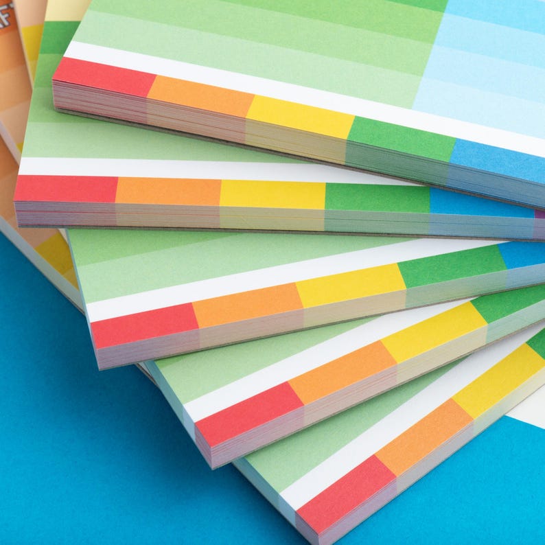 Puede incluir: Una pila de cuadernos de papel de colores con rayas verdes, blancas, naranjas, amarillas, rojas y azules. Los cuadernos est&aacute;n dispuestos en forma de abanico sobre una superficie azul.
