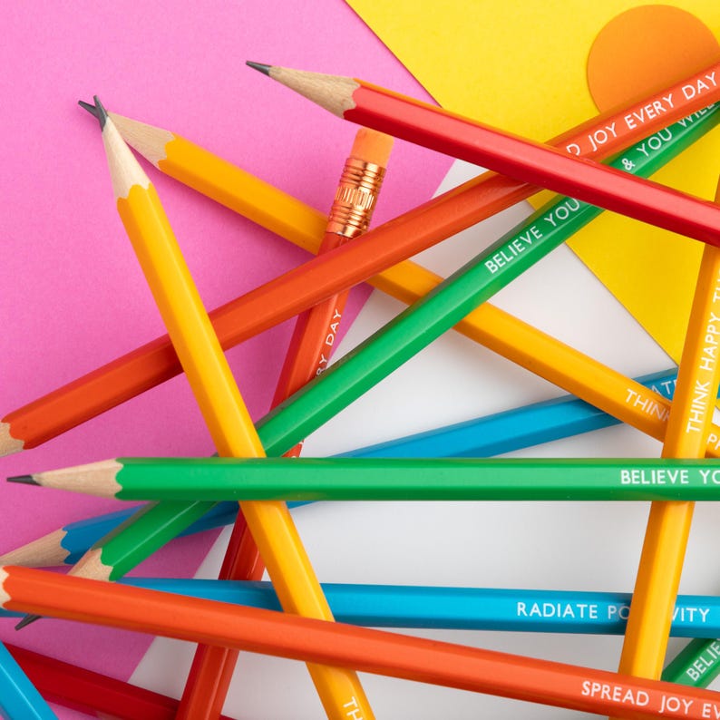 Pu&ograve; includere: Una collezione di matite colorate con testo, tra cui "Believe You" e "Spread Joy Every Day". Le matite sono in vari colori: giallo, arancione, verde e blu. Le matite sono disposte su uno sfondo di carta rosa, bianca e gialla.