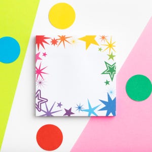 May include: A square white notepad with a colourful star border in shades of red, orange, yellow, green, blue, and purple. The notepad is surrounded by colourful polka dots on a white, pink, and green background. Stationery.