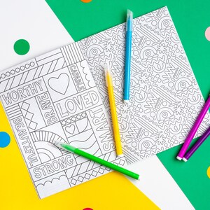 Puede incluir: Un libro para colorear con un dise&ntilde;o en blanco y negro que incluye palabras como "digno", "valiente", "amado", "capaz" y "inteligente". El libro est&aacute; abierto sobre una superficie amarilla y hay cinco marcadores de colores junto a &eacute;l.