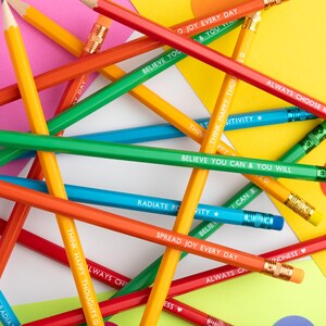 Pu&ograve; includere: Una collezione di matite colorate con frasi motivazionali come "Believe You Can & You Will" e "Spread Joy Every Day". Le matite sono di vari colori, tra cui rosso, arancione, giallo, verde e blu, con fasce metalliche dorate e gomme rosa.