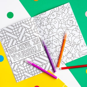 Puede incluir: Un libro para colorear con un patr&oacute;n geom&eacute;trico en blanco y negro en un lado y la frase "Cree en ti mismo" en el otro lado. El libro est&aacute; abierto y hay cuatro marcadores de colores encima.