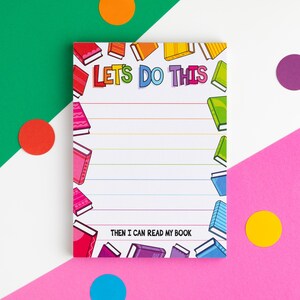 May include: A rectangular notepad featuring the text LET'S DO THIS at the top and THEN I CAN READ MY BOOK at the bottom. The page has horizontal lines and is decorated with colourful illustrations of books along the left and right borders.