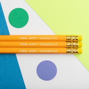 Puede incluir: Tres lápices amarillos con la frase "THINK HAPPY THOUGHTS" impresa. Los lápices tienen gomas de borrar amarillas y bandas metálicas doradas. El fondo es un diseño geométrico con formas verdes, azules, blancas y amarillas.
