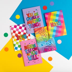 Puede incluir: Cinco cuadernos coloridos con diferentes diseños y mensajes inspiradores. Los cuadernos tienen rayas arcoíris, patrones a cuadros y un diseño con útiles escolares. Los mensajes en los cuadernos incluyen "You are capable of amazing things", "Ideas", "Believe you can and you will", y "Notes".
