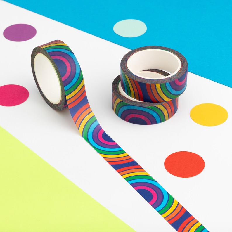 Puede incluir: Tres rollos de cinta adhesiva decorativa con un dise&ntilde;o retro de arco&iacute;ris. Un rollo est&aacute; desenrollado, revelando un patr&oacute;n de arco colorido. La cinta est&aacute; sobre una superficie blanca con lunares coloridos y un fondo azul, verde y amarillo.