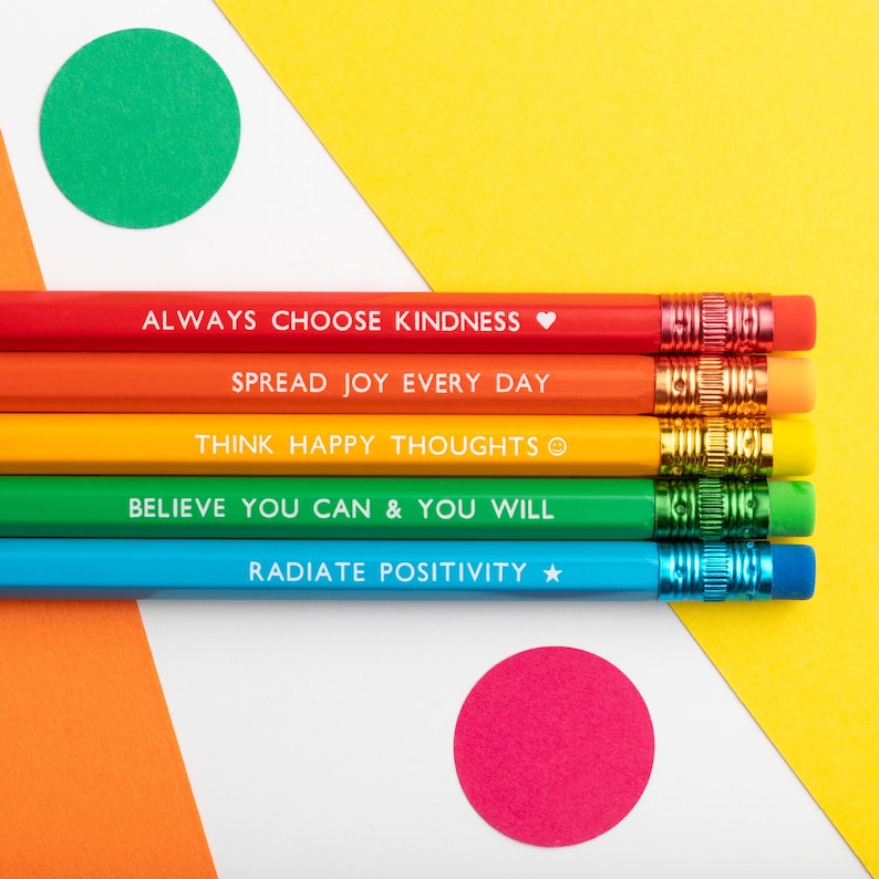 Pu&ograve; includere: Primo piano di cinque matite colorate arcobaleno con frasi motivazionali in testo bianco. Le matite sono disposte orizzontalmente, con il testo: "ALWAYS CHOOSE KINDNESS," "SPREAD JOY EVERY DAY," "THINK HAPPY THOUGHTS," "BELIEVE YOU CAN & YOU WILL," e "RADIATE POSITIVITY."