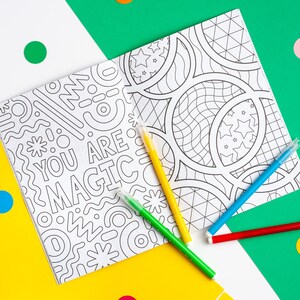 Puede incluir: Dos p&aacute;ginas para colorear con ilustraciones en blanco y negro. Una p&aacute;gina presenta un patr&oacute;n geom&eacute;trico con el texto "You Are Magic". La otra p&aacute;gina presenta un patr&oacute;n de c&iacute;rculos y estrellas. Cuatro marcadores de colores est&aacute;n encima de las p&aacute;ginas.
