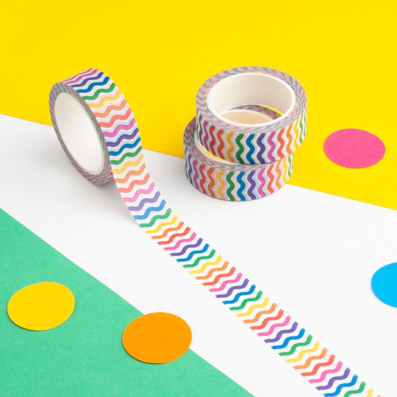 May include: A roll of colorful washi tape with a wavy pattern. The tape is unrolled and laying on a white surface with a yellow surface in the background. There are three colorful circles on the surface.