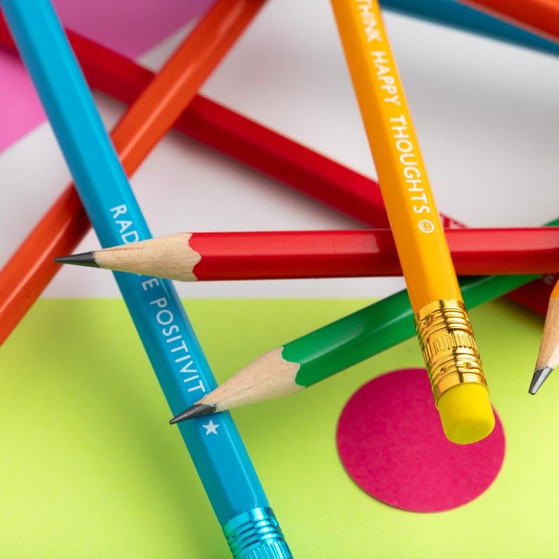 Pu&ograve; includere: Primo piano di matite colorate vivaci, tra cui blu, arancione, rosso e verde, con i testi "RAD E POSITIVITY" e "THINK HAPPY THOUGHTS". Le matite sono disposte su uno sfondo di carta rosa, verde e bianca.
