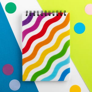 May include: A spiral-bound notebook with a white cover featuring a wavy rainbow pattern. The notebook's design includes stripes of purple, red, orange, yellow, green, and blue. The background has colorful confetti and geometric shapes.