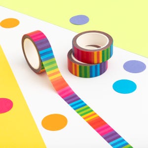 May include: Washi tape rolls with a rainbow striped design. One roll is unrolled, showing a pattern of horizontal stripes. Two rolls are stacked, displaying the same vibrant design. The background includes colourful confetti.