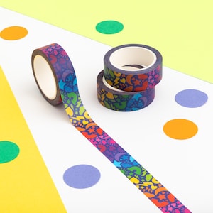 May include: Decorative washi tape rolls with a rainbow pattern of stars, hearts, lightning bolts, and question marks. One roll is unrolled, showing the colorful design. Two additional rolls are stacked on a white surface with colorful confetti.