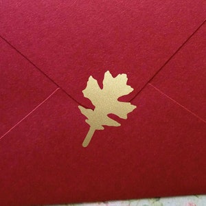Pode incluir: Um envelope vermelho com um adorno dourado em forma de folha. O envelope está fechado e tem uma superfície texturizada. A folha está centrada na frente do envelope.