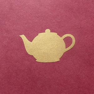 May include: A gold-colored teapot cutout with a rounded body, spout, and handle. The teapot is set against a deep red background. The cutout is a simple, elegant design, suitable for crafts or decorative purposes.