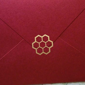 May include: A deep red envelope with a gold honeycomb design. The envelope is sealed and has a textured surface. The background is a floral pattern on a wooden surface. The envelope is a stationery item.