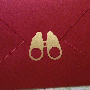 May include: A red envelope with a gold-colored binoculars design. The envelope is rectangular and has a matte finish. The binoculars are centered on the front of the envelope.