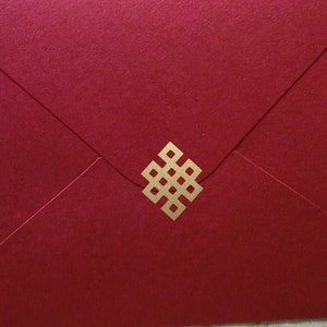 Puede incluir: Un sobre rojo intenso con un emblema decorativo dorado. El sobre es rectangular y tiene un cierre de solapa. El fondo presenta un estampado floral sobre una superficie de madera clara.