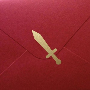 Könnte beinhalten: Ein roter Umschlag mit einem goldenen schwertförmigen Emblem. Der Umschlag ist rechteckig und versiegelt, mit dem Emblem auf der Vorderseite. Der Hintergrund ist ein Blumenmuster.