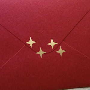Può includere: Una busta rossa con accenti a forma di stella dorata. La busta è chiusa e pronta per essere spedita.