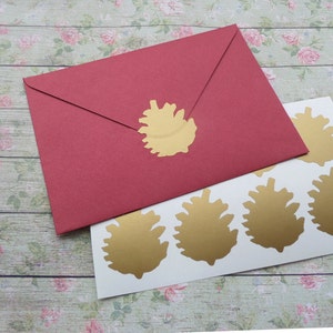 Puede incluir: Un sobre rojo sellado con una pegatina dorada de piña. Varias pegatinas doradas de piña están sobre una hoja blanca. Los artículos están sobre una superficie con estampado floral. La imagen muestra artículos de papelería y artesanía.