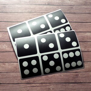 May include: A set of nine black and white dice stickers with different numbers of dots on each die. The stickers are arranged in a grid pattern on a sheet of paper.