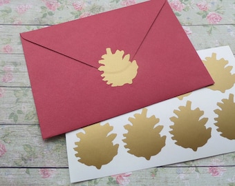 May include: A red envelope sealed with a gold pinecone sticker. Several gold pinecone stickers are on a white sheet. The items are on a floral patterned surface. The image showcases stationery and craft supplies.