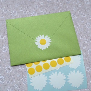 Puede incluir: Un sobre verde claro con una pegatina de margarita, junto con una hoja de pegatinas de c&iacute;rculo amarillo y una hoja de pegatinas de margarita blanca con centros amarillos. El fondo es un estampado floral.