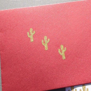 Op de afbeelding: Een rode envelop met drie gouden glitter cactusontwerpen. Een vel witte stickers met meerdere gouden glitter cactusontwerpen is gedeeltelijk zichtbaar. De afbeelding is goed belicht en toont de producten in detail.
