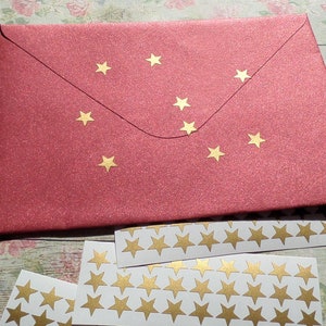 Op de afbeelding: Een rode envelop met gouden ster stickers. Er zijn meerdere vellen wit papier met gouden ster stickers.