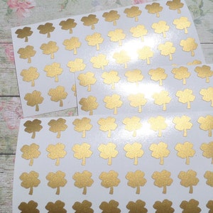 Könnte beinhalten: Blätter mit goldenen Kleeblatt-Aufklebern auf weißem Hintergrund. Jedes Blatt ist mit mehreren vierblättrigen Kleeblatt-Designs gefüllt. Die Aufkleber sind in ordentlichen Reihen und Spalten angeordnet und bilden ein Muster. Die goldene Farbe sorgt für ein glänzendes, metallisches Aussehen.