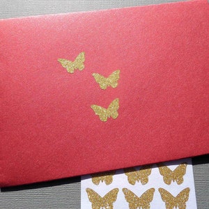 Op de afbeelding: Een rode envelop met gouden glitter vlinder stickers. De envelop ligt op een grijs oppervlak. Er zijn meer gouden glitter vlinder stickers op een wit vel papier onder de envelop.