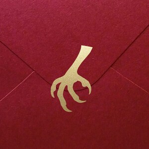 Könnte beinhalten: Ein roter Umschlag mit einem goldenen Krallen-Design. Der Umschlag besteht aus strukturiertem Papier und ist versiegelt. Der Hintergrund ist ein Blumenmuster auf einer Holzoberfläche.